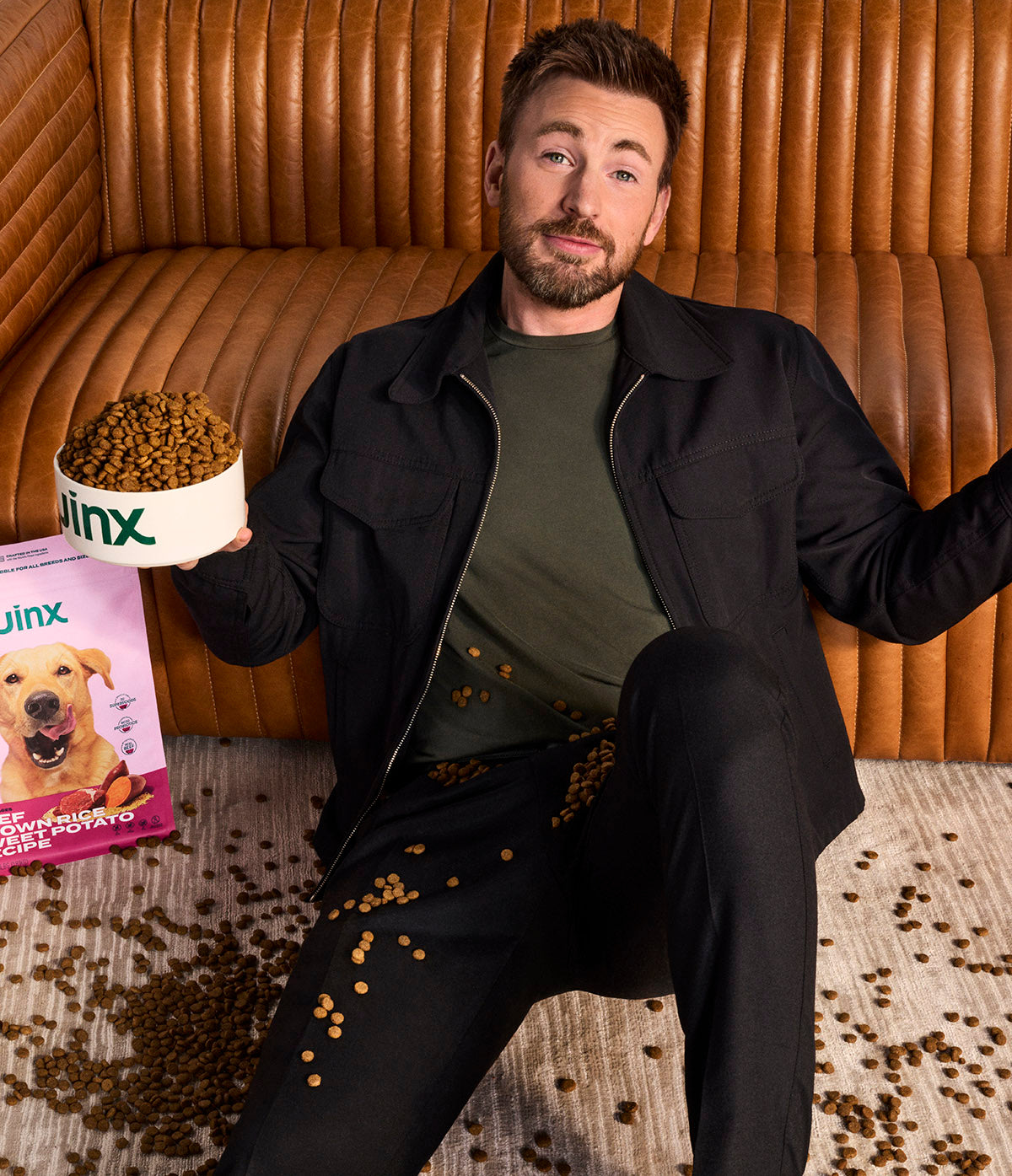 an image of Chris Evans sitting on the ground shrugging, covered in Jinx kibble.