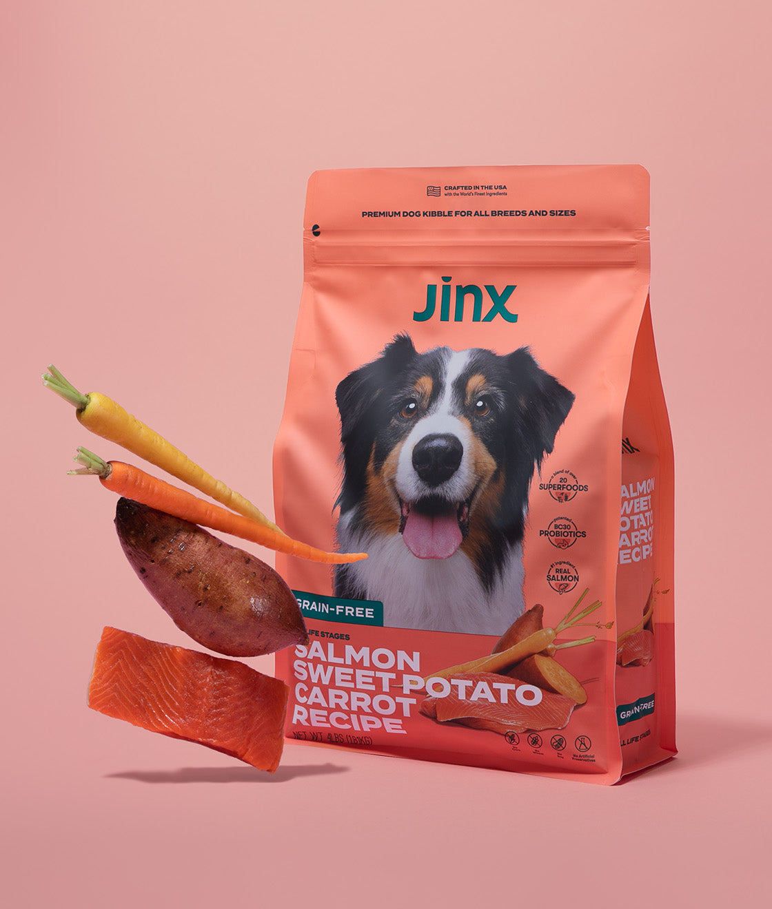 Salmon, sweet potato and carrot Jinx dog kibble product packaging, with those ingredients hovering in front of it on a pink background.