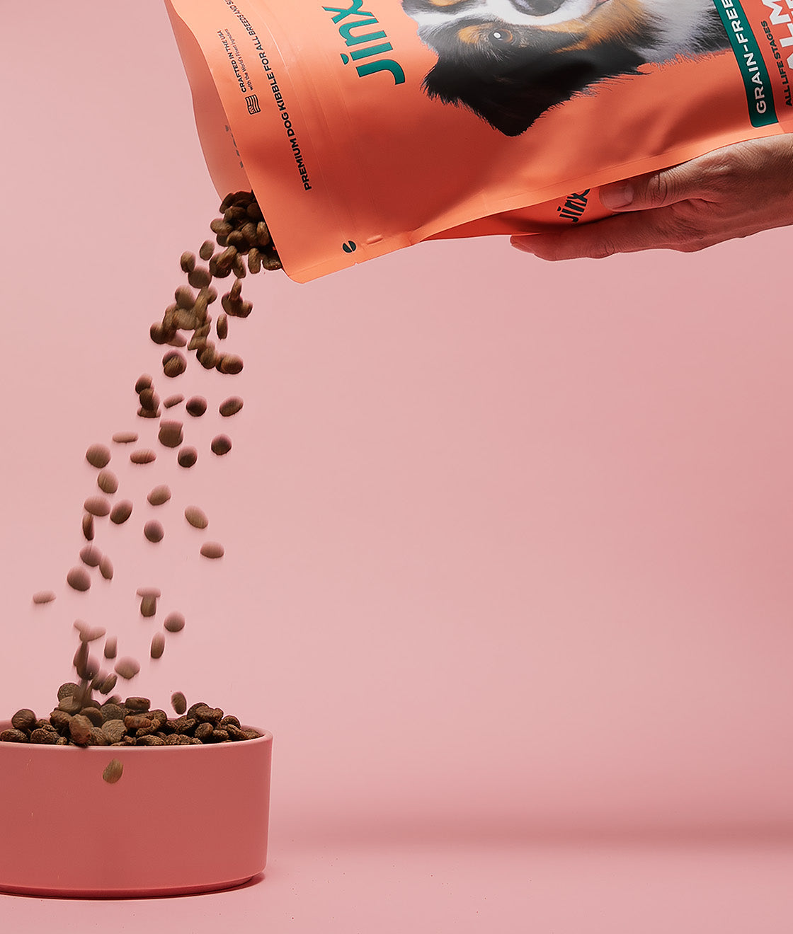 A hand holding a bag of Jinx dog kibble and filling up a pink dog bowl with a pink background