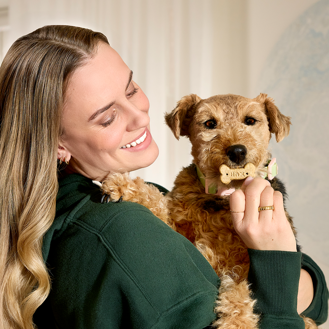 Woman holding a dog with a bone-shaped treat in its mouth, both smiling.