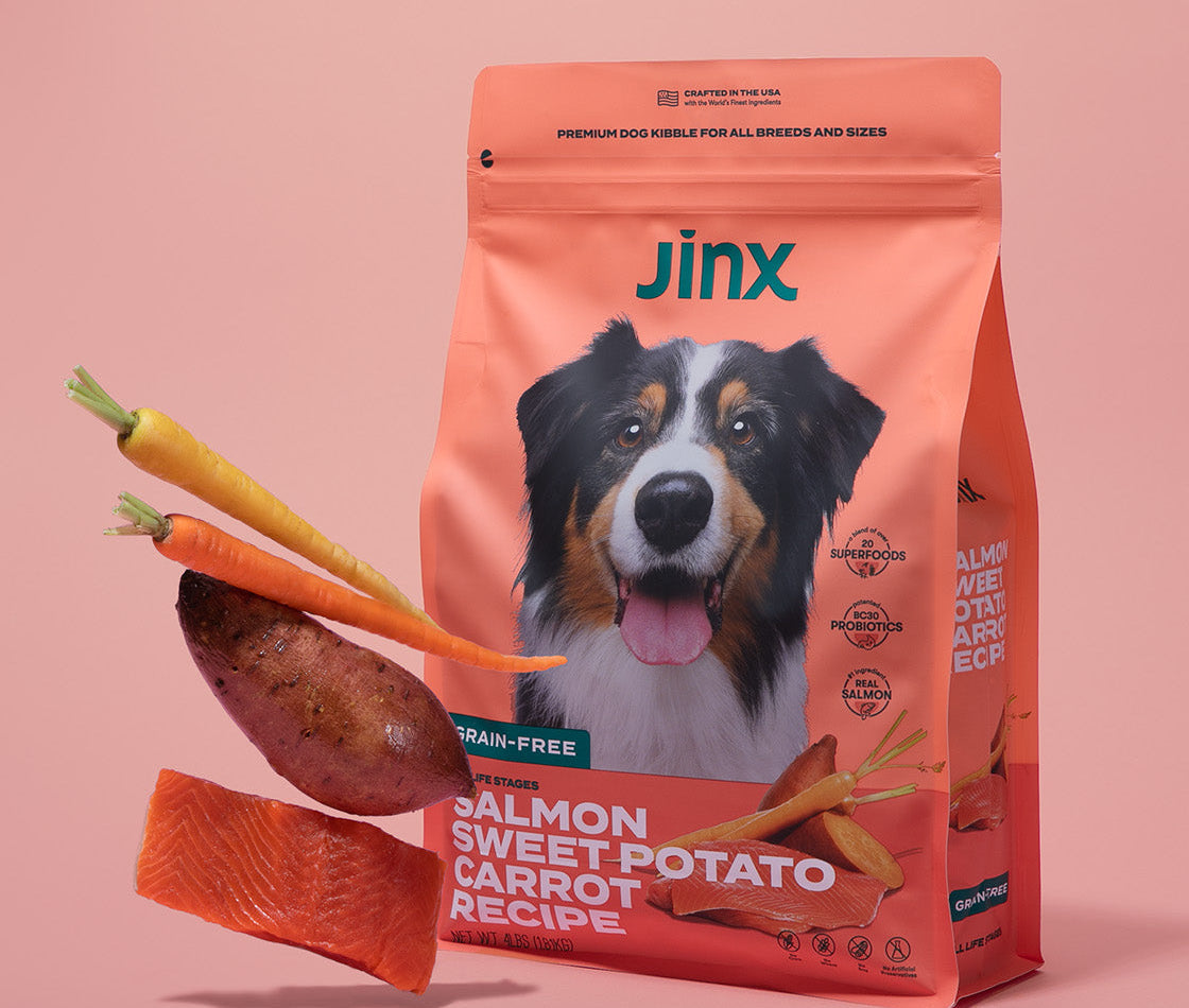 Salmon, sweet potato and carrot Jinx dog kibble product packaging, with those ingredients hovering in front of it on a pink background.